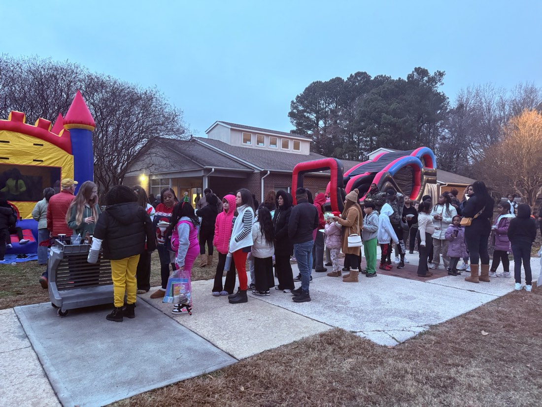 A crowd in winter coats on the sidewalk at dusk, with bounce houses behind them.
